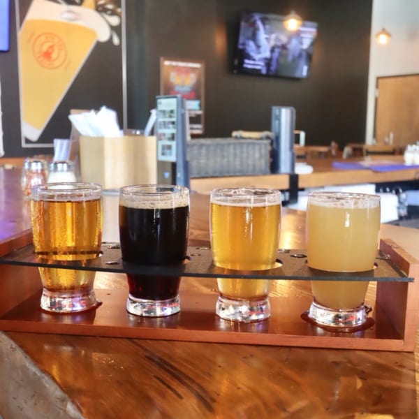 Four beer samples on a wooden flight paddle on a bar counter with a beer mural in the background.