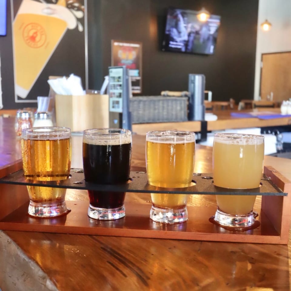 Four beer samples on a wooden flight paddle on a bar counter with a beer mural in the background.