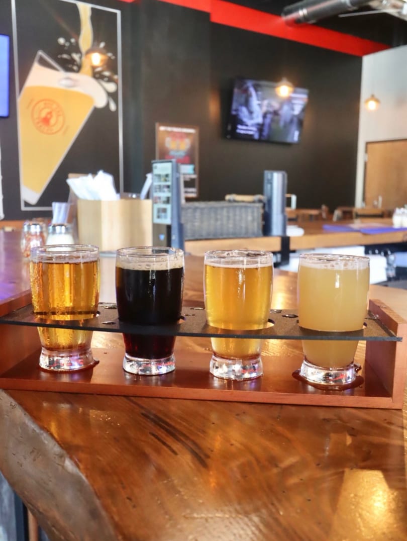 Four beer samples on a wooden flight paddle on a bar counter with a beer mural in the background.