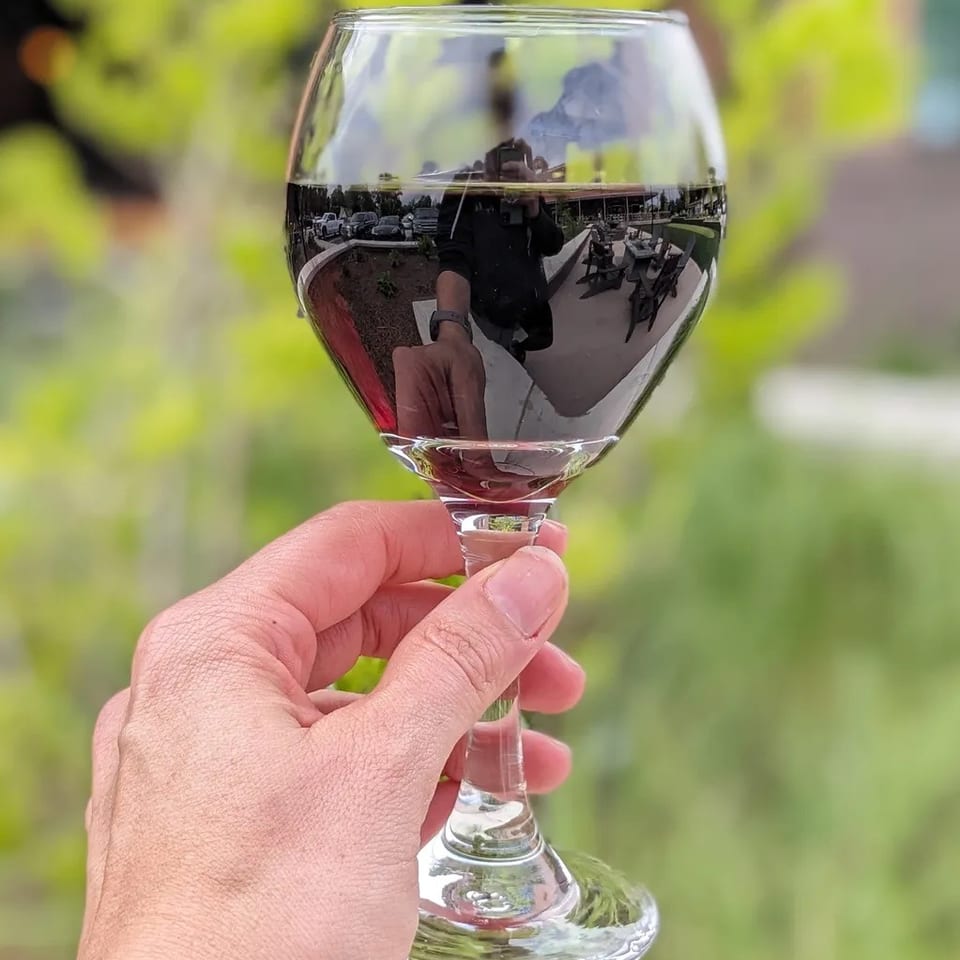 Hand holding a red wine glass, reflecting a person, outdoor patio, and cars, against green foliage.