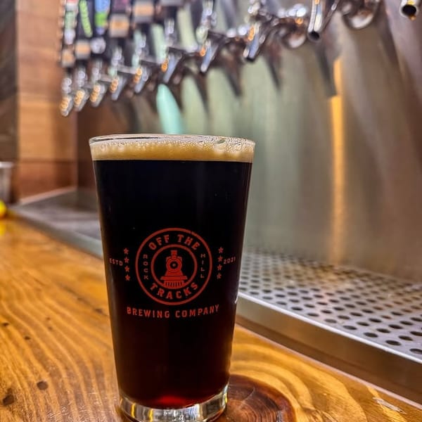 A dark beer in a branded glass sits on a wooden bar counter with beer taps in the background.