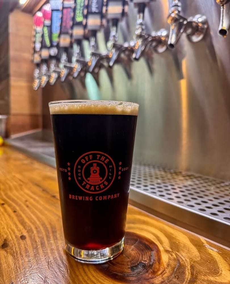 A dark beer in a branded glass sits on a wooden bar counter with beer taps in the background.