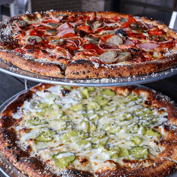 Two pizzas on metal stands, one with red sauce and meats, the other with white sauce and green peppers.