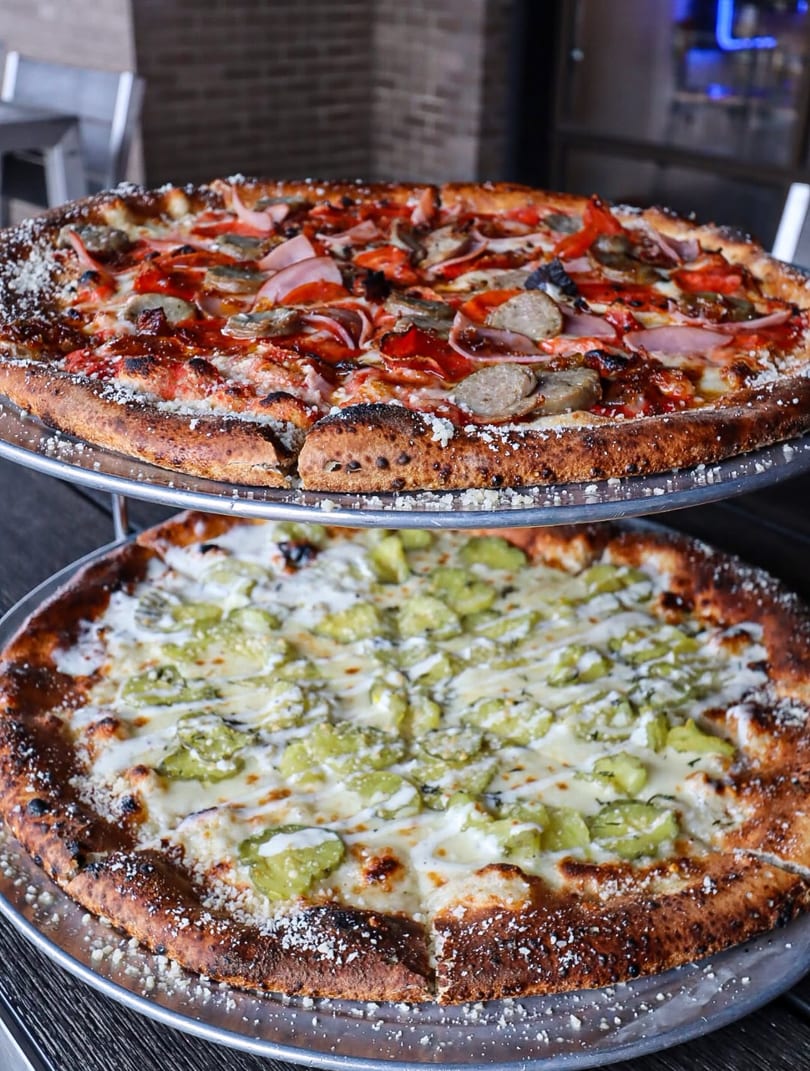 Two pizzas on metal stands, one with red sauce and meats, the other with white sauce and green peppers.