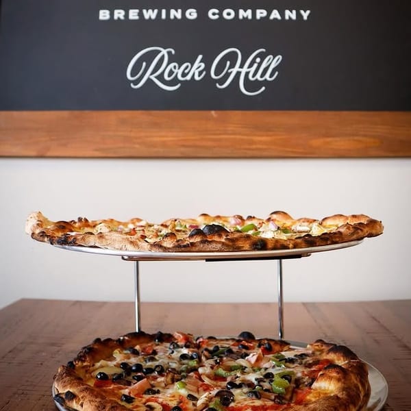 Two pizzas, one on a stand, with a "Off the Tracks Brewing Company Rock Hill" sign in the background.