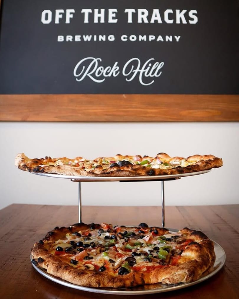 Two pizzas, one on a stand, with a "Off the Tracks Brewing Company Rock Hill" sign in the background.