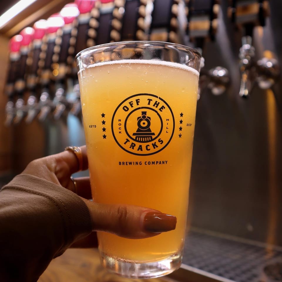 Hand holding a glass of Off The Tracks Brewing Company beer with taps in the background.