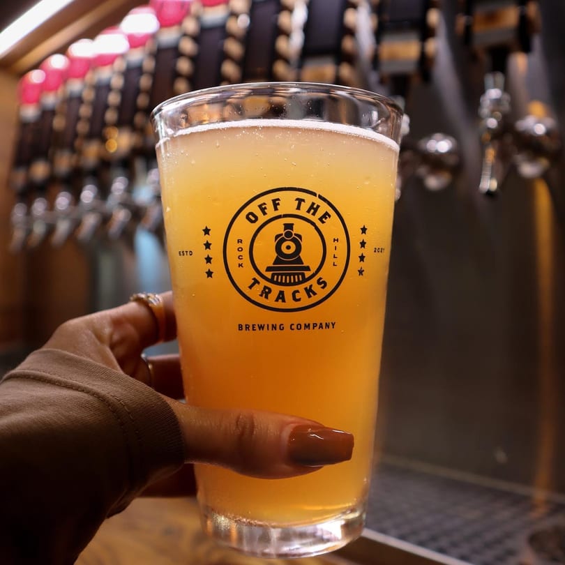 Hand holding a glass of Off The Tracks Brewing Company beer with taps in the background.