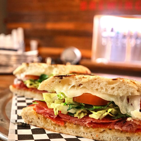 Two grilled Italian subs with melted cheese, salami, lettuce, and tomato on a checkered paper.
