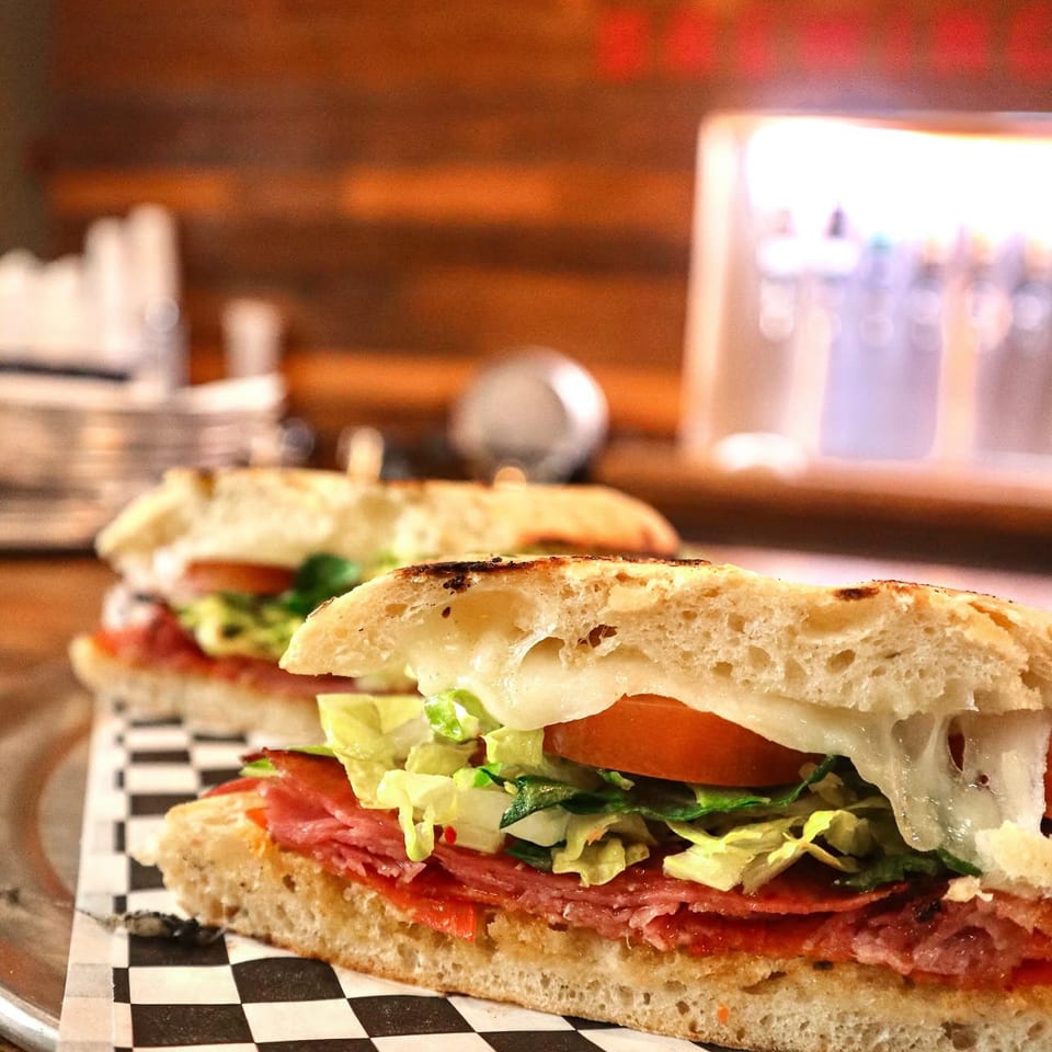 Two grilled Italian subs with melted cheese, salami, lettuce, and tomato on a checkered paper.
