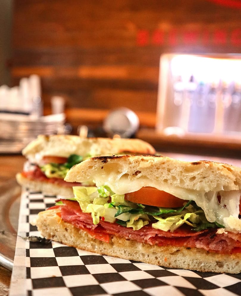 Two grilled Italian subs with melted cheese, salami, lettuce, and tomato on a checkered paper.