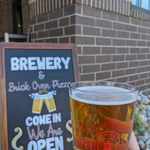 Hand holding a beer glass with "Off The Tracks Brewing Company" logo in front of a "Brewery & Brick Oven Pizza" sign.