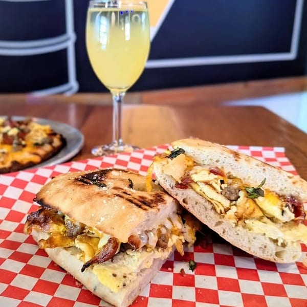 Grilled breakfast sandwich with egg, bacon, and cheese, served with a drink and pizza.