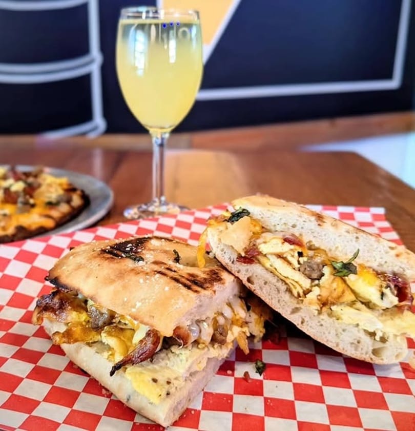 Grilled breakfast sandwich with egg, bacon, and cheese, served with a drink and pizza.