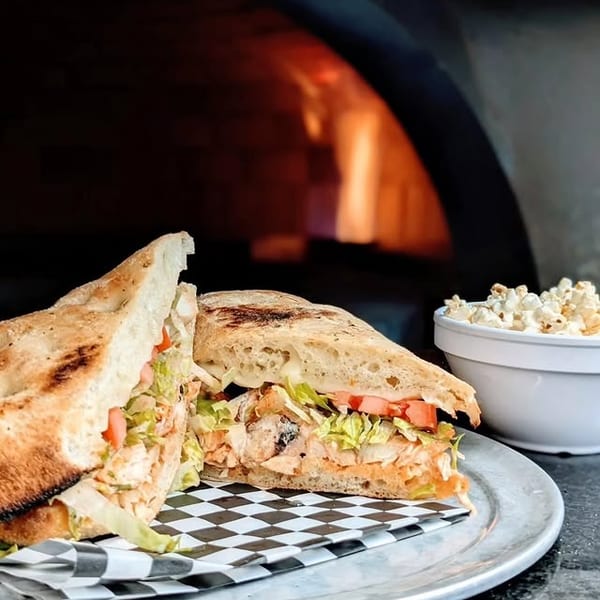 Chicken sandwich with lettuce and tomato on a plate, with a wood-fired oven and popcorn in the background.