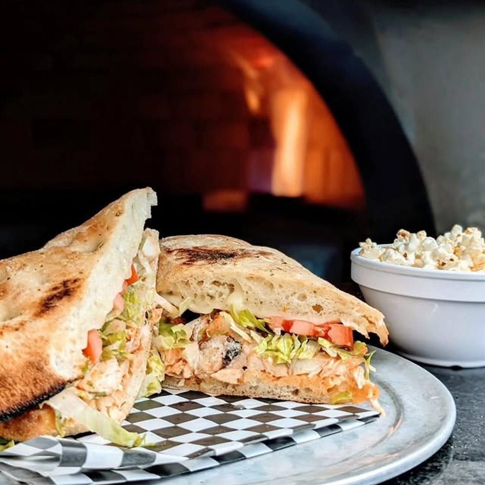 Chicken sandwich with lettuce and tomato on a plate, with a wood-fired oven and popcorn in the background.