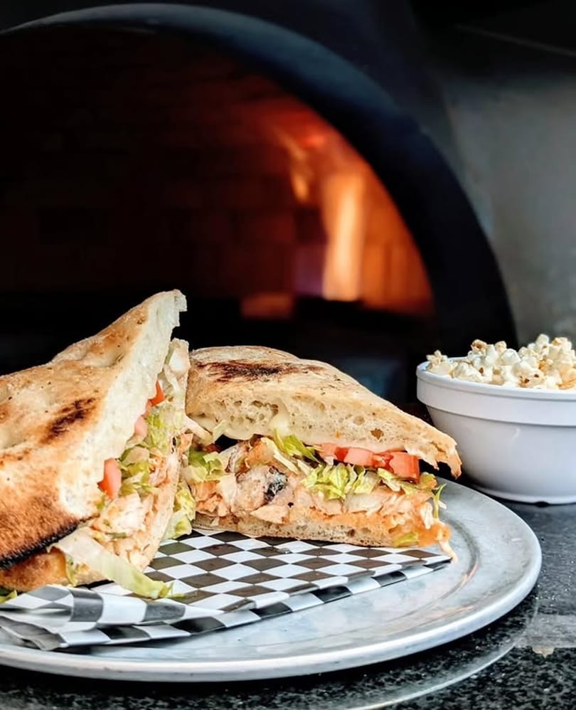 Chicken sandwich with lettuce and tomato on a plate, with a wood-fired oven and popcorn in the background.