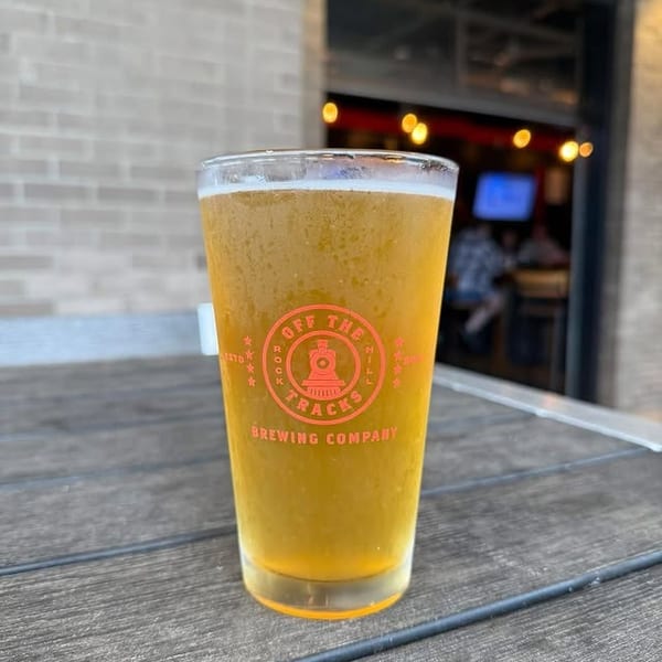 A pint glass of golden beer with the "Off The Tracks Brewing Company" logo sits on a wooden table.