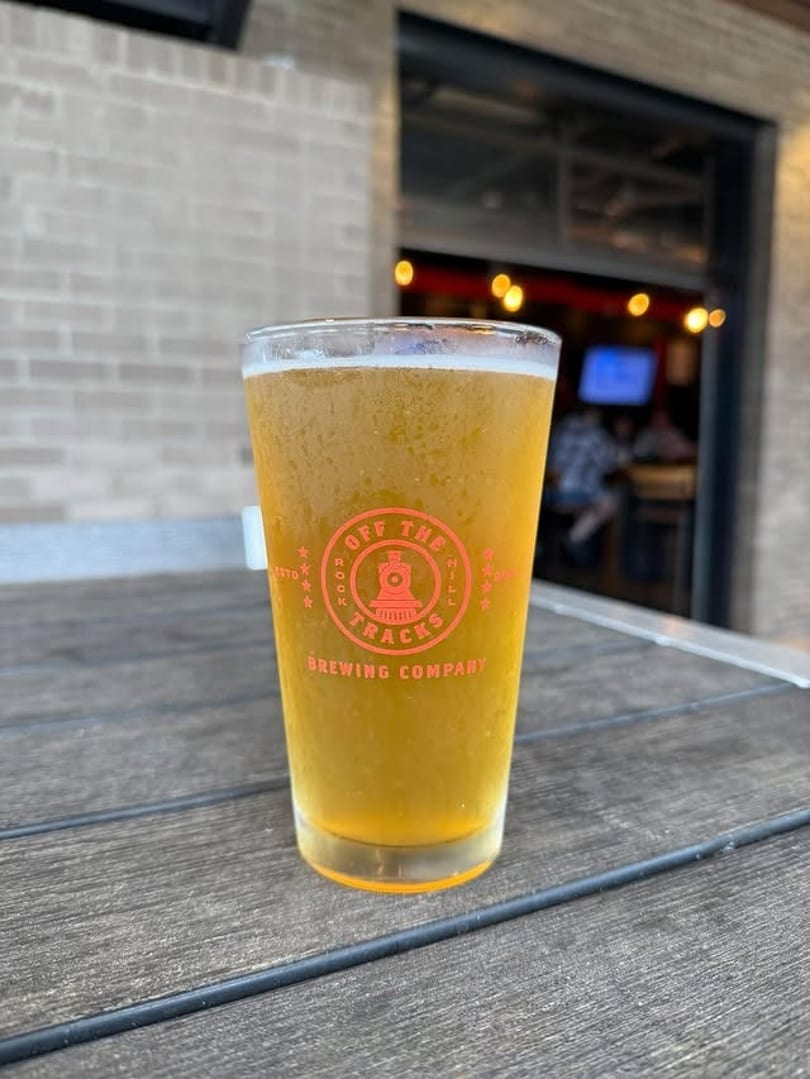 A pint glass of golden beer with the "Off The Tracks Brewing Company" logo sits on a wooden table.
