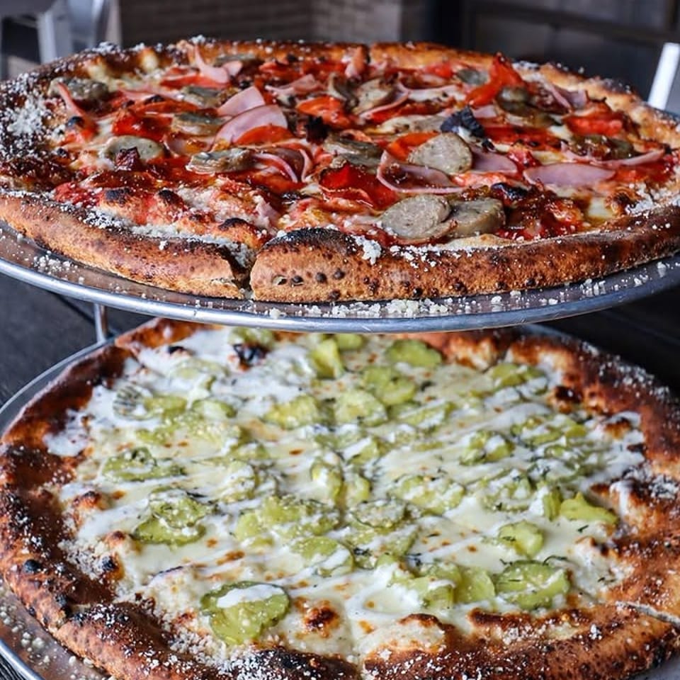 Two pizzas on metal stands, one with meat and red sauce, the other with white sauce and pickles.