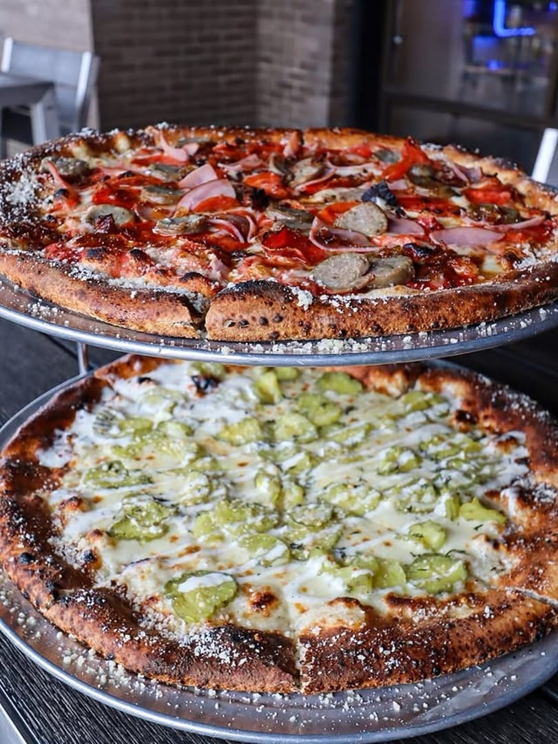 Two pizzas on metal stands, one with meat and red sauce, the other with white sauce and pickles.