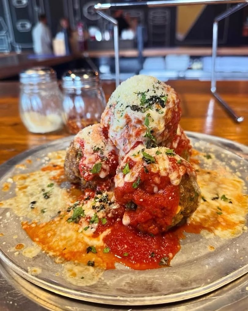 Four large meatballs covered in marinara sauce, melted cheese, and fresh parsley on a metal plate.