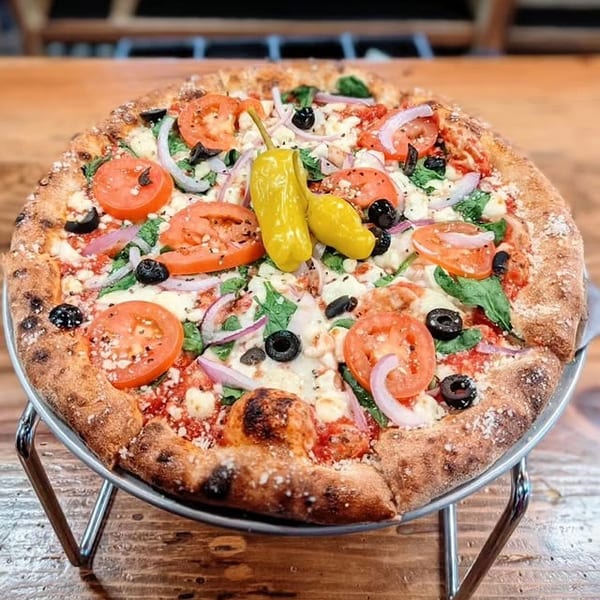 A whole pizza with tomatoes, olives, red onions, spinach, and two yellow peppers on a metal stand.