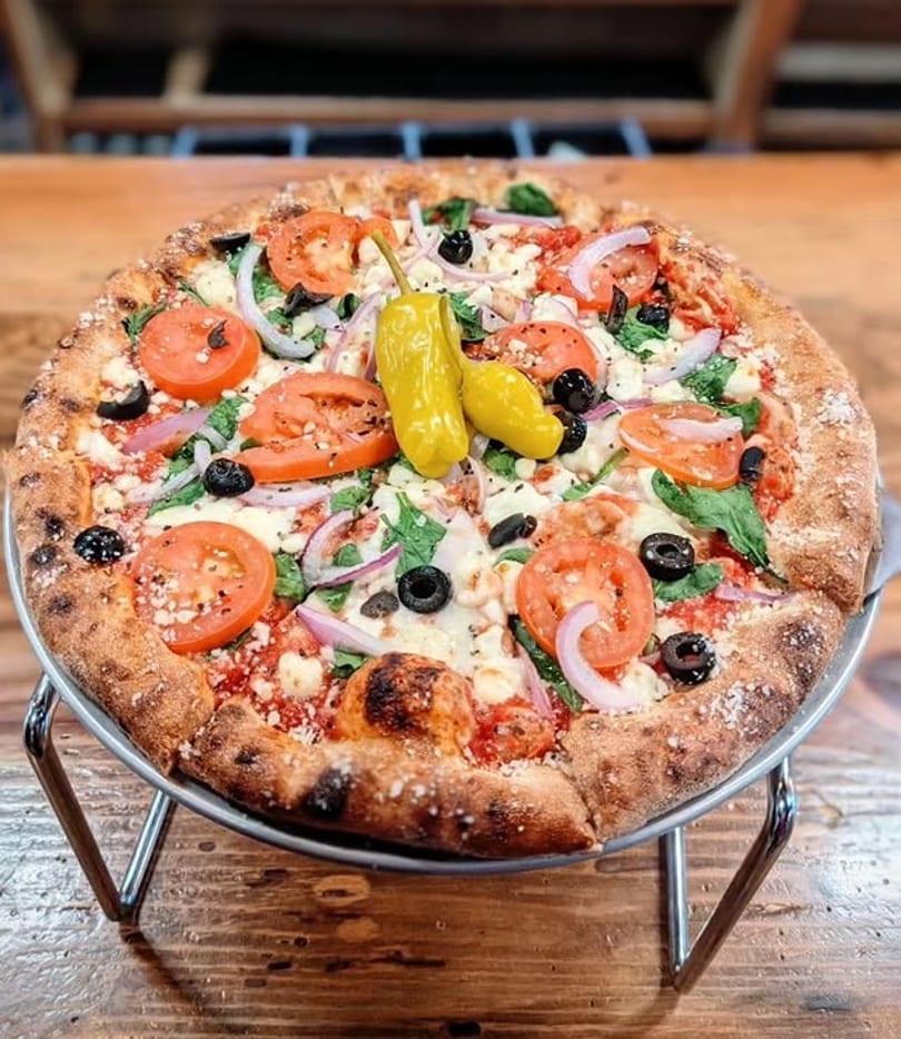 A whole pizza with tomatoes, olives, red onions, spinach, and two yellow peppers on a metal stand.