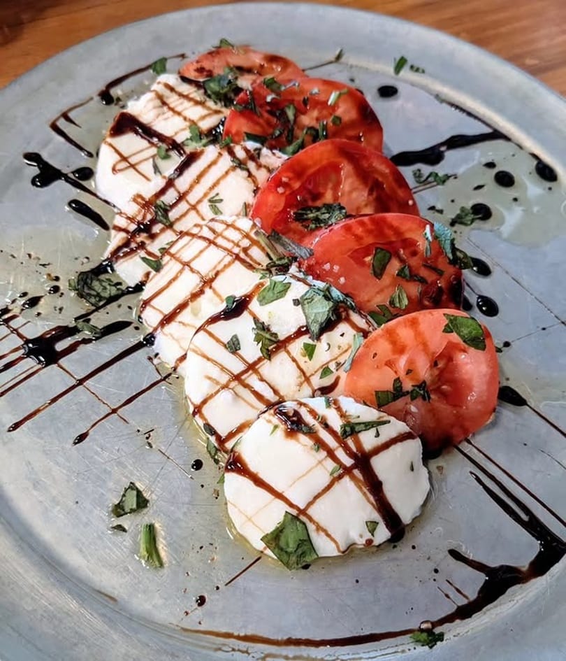 Caprese salad with sliced mozzarella, tomatoes, basil, and balsamic glaze on a silver plate.