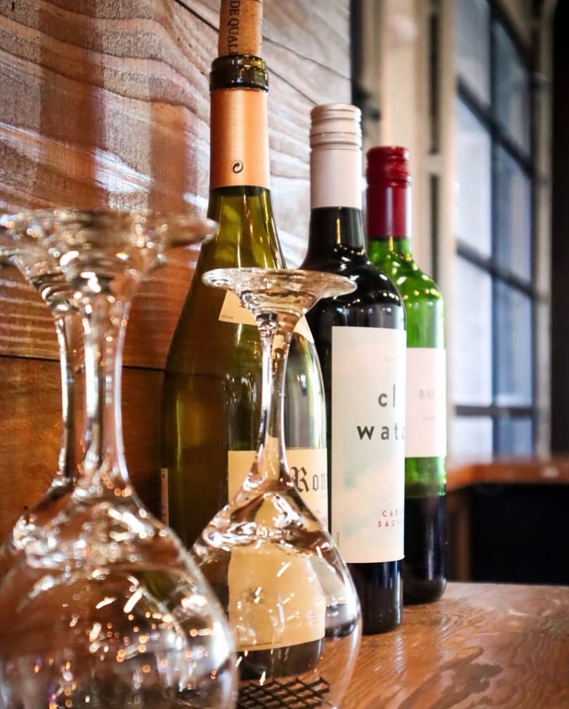 Wine bottles and inverted wine glasses are displayed on a wooden surface against a wood-paneled wall.