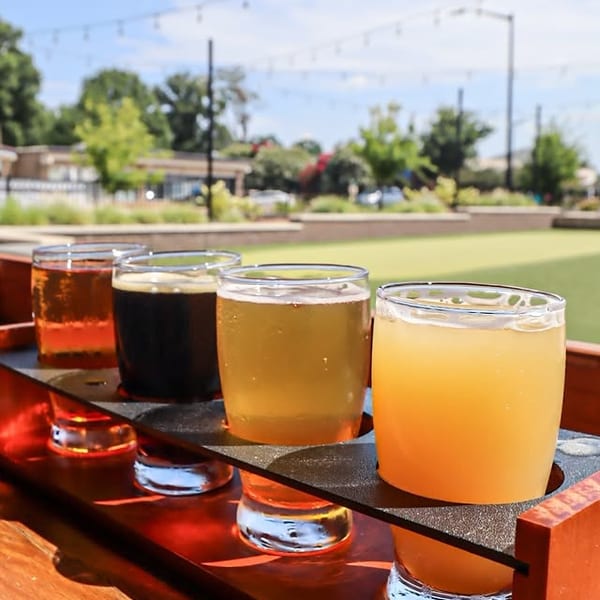 A flight of four craft beers on a wooden tray, outdoors on a sunny day.