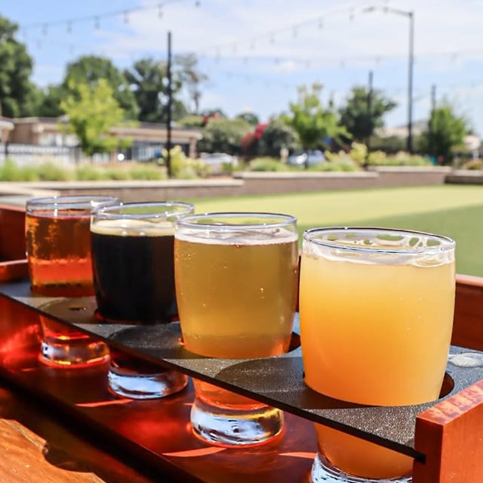 A flight of four craft beers on a wooden tray, outdoors on a sunny day.