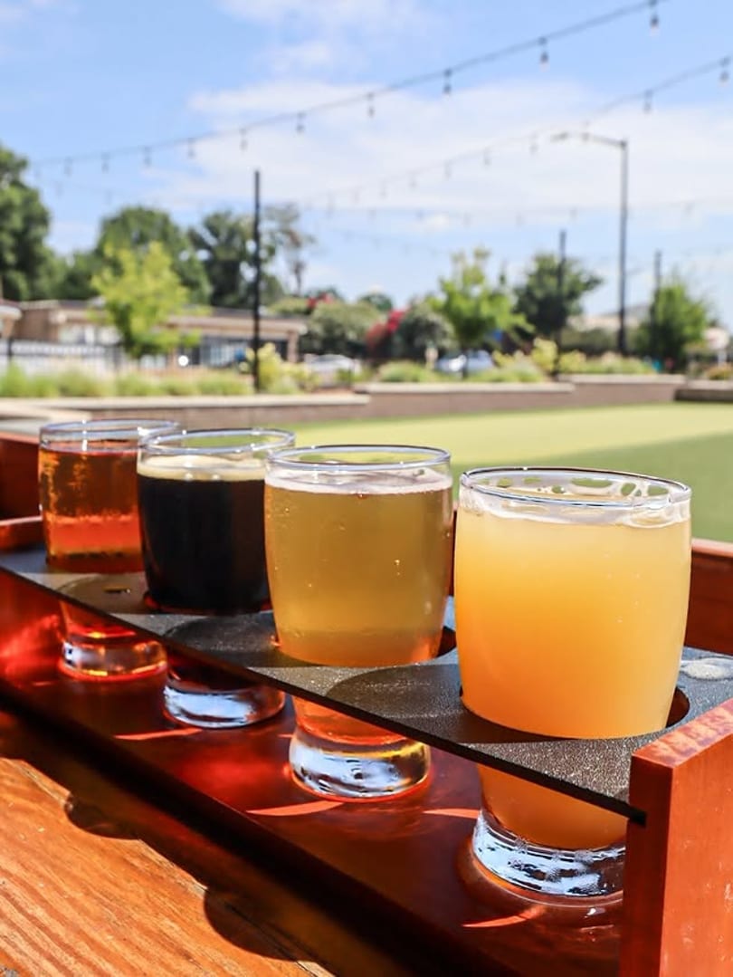 A flight of four craft beers on a wooden tray, outdoors on a sunny day.